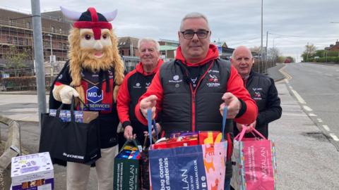Skinn holds a bag for life crammed with easter eggs. Two other men behind him also stand with bags as does the team mascot in a cartoon viking mask.