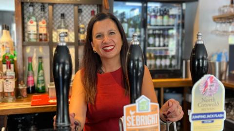 Emma Gibbon is pictured in the centre of the frame, behind the bar. She has dark hair and is wearing red top and is smiling from behind the bar taps. Around her is a typical bar scene, spirits on the left of her at the back and on the right is a fridge full of bottled beers and ciders. The photo itself is in portrait mode making her and the taps in front of her, the focus, with the rest of the pub slightly blurred.