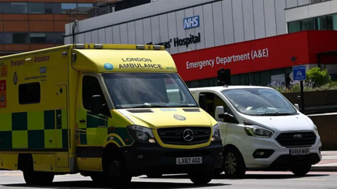 A yellow ambulance with the words London Ambulance parked next to a white van outside a hospital emergency department 