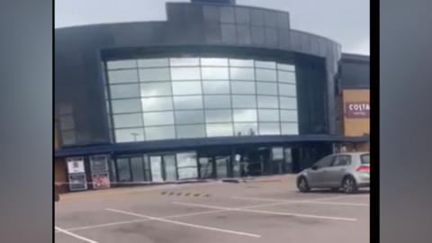 A still taken from a video, which is slightly blurry. It shows the front of the cinema with glass doors damaged, and red hazard tape across the car park. Above the main doors are glass mirrored panels.