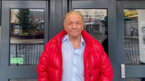 Arthur Holland is stood outside the doors to the Emporium in Coalville. He is wearing a red puffer jacket and a blue shirt. 