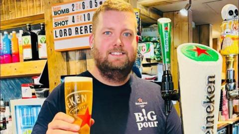 Tom stands behind a bar and holding up a tall pint glass of Heineken beer toward the camera. The glass is filled with golden lager and has a thick white head at the top. He is wearing a dark navy or black T-shirt with the words “BIG pour” and a small Heineken logo printed on the chest.