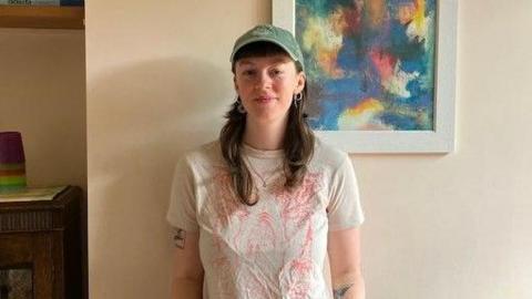 A woman standing against a cream wall. She is wearing a white top and has tattoos on her arms. She is smiling and wearing a baseball cap.