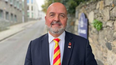 A picture of a man with no hair and a grey beard. He is wearing a navy suit jacket with a white shirt and red and yellow striped tie.