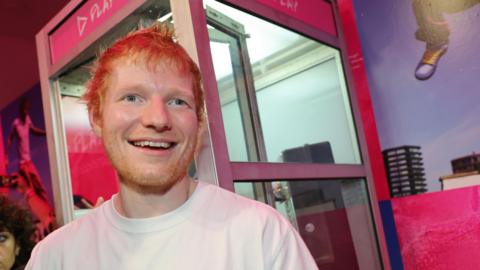 Ed Sheeran smiles in front of a pink phone box themed around his album title, Play.