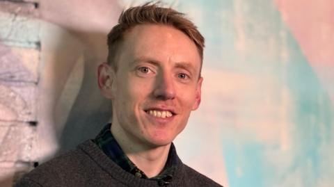A clean-cut man in his thirties is looking straight at the camera and smiling. We can just see a dark sweater and a black shirt. He's in front of a wall painted a colourful mix of lights pinks and blues 
