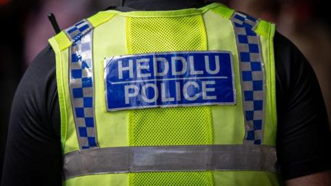 A police officer wearing a black t-short, with their back to the camera. Their yellow vest reads 'Heddlu, Police' in a blue panel. 