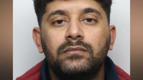 A custody photo of a man with dark short hair and dark facial hair and brown eyes wearing a red and blue top. He is sat in front of a grey background.