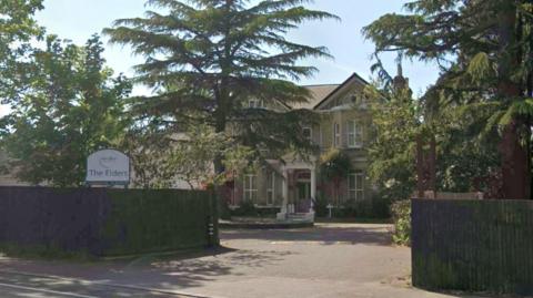 The image shows the outside of a care home, with a sign on the left hand side reading "The Elders". There are many trees surrounding the building which are placed behind wooden fences. The house in the background is large with many windows.