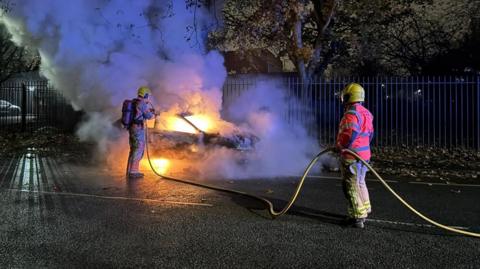 Two firefighters hold a yellow hose as they put out a car on fire on a road at night in Salford