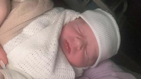 The baby is wearing a white hat and is wrapped in a white blanket. His eyes are closed. He is being held by someone with blue and pink nails.