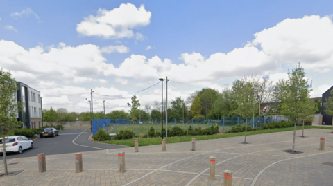 A large paved area with bollards and trees behind it, with a small road on the left hand side