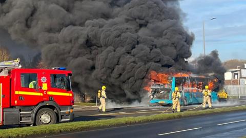 A red fire engine is parked on a dual carriageway ahead of a single-decker turquoise bus which is in the grip of a massive fire. Thick clouds of black smoke billow into the air and orange flames are visible from the bus windows. Three firefighters can be seen standing close to the bus, spraying it with fire-hoses. 