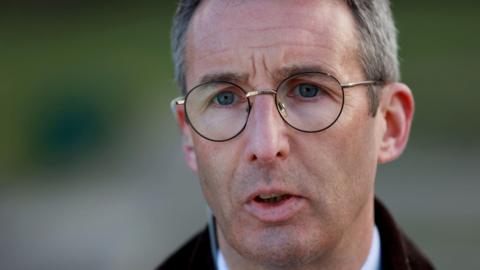 Andrew Muir is speaking to the press wearing a brown jacket and has glasses on. He has short white/grey hair.