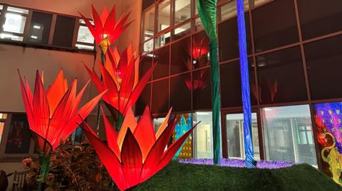 Five tall flower sculptures which are lit up in deep red hues at night outside the hospital, where lights are on in the windows. 