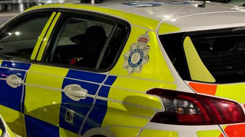 A close-up of a police car with their emblem showing Humberside Police. There are blue and bright-yellow squares on the paintwork and the car has tinted windows.