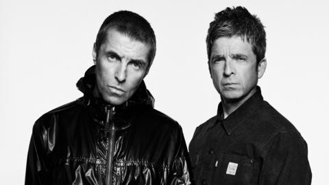 A black and white photo of two men looking into a camera. Liam, on the left, is wearing a shiny jacket with a zip up the middle and has sort shaved hair. Noel is to his left and is wearing a buttoned-up jacket and also has short hair but his is styled.