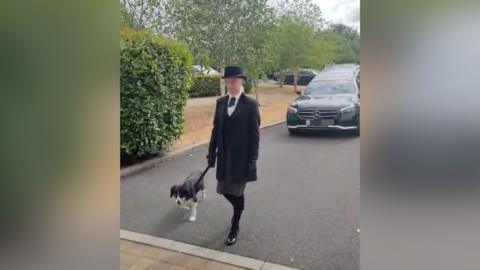 A woman in a black suit with a white shirt, black shoes, gloves and black bowler hat is walking in front of a hearse with a dog