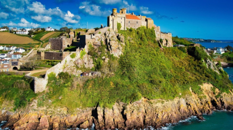 The image shows a large medieval castle perched on a steep, rocky headland beside the sea. The stone walls and towers stretch across the top of the cliff, with patches of greenery climbing over the ruins. Below the fortifications, jagged rocks drop into the water. Farmland and houses sit further inland behind the castle under a bright blue sky with scattered clouds.