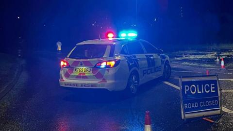 A police car parked at an angle across a sliproad blocks the entrance to a road. A "POLICE ROAD CLOSED" sign is positioned in the foreground.