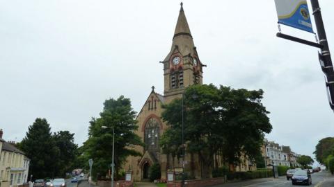 A view of the Hornsea United Reformed Church, which is built from yellow brick and has a pointed steeple containing a clock