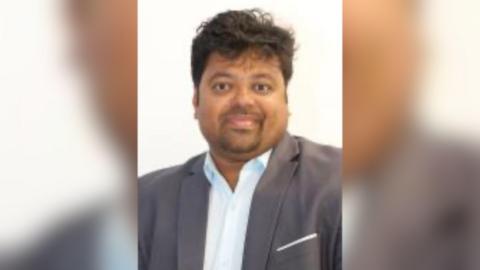 Portrait image of Leedo George. He is looking into camera and has dark hair, a short goatee beard and is wearing a suit jacket and shirt with the top button undone.