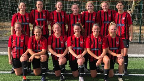 A women's football team in a traditional pose of two rows with the women at the back standing and the women in the front crouching neatly. They are dressed in red and black striped kits.