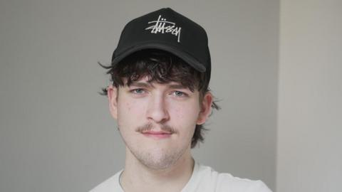 Hamish Wilson pictured at the University of Southampton where he is a student. Hamish is wearing a black cap and has dark tousled hair. He is looking to camera and is wearing a white t-shirt.