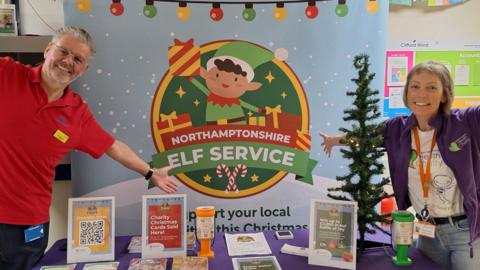 A man in a red polo and a woman in a purple fleece stand with their arms out invitingly in front a large "Northamptonshire Elf Service" sign with a green elf dishing out presents on display. There are leaflets and framed information on a table.