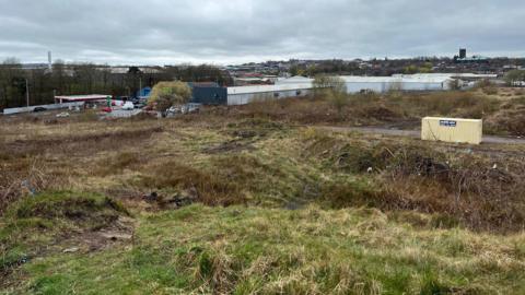 An area of land behind a set of industrial units with a petrol station and Tunstall town centre in the distance.