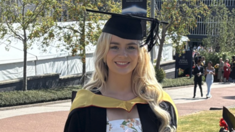 A woman with long blonde hair is wearing black graduation clothing and a white dress including floral designs. A grey building and people are in the distance.