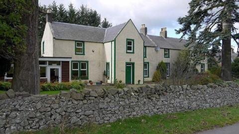 A white house with green door and window frames surrounded by trees with a dry stone wall in front of it