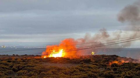 A fire at a colliery bing in Methil. The flames are orange with a plume of dark grey smoke coming off it. Green shrubbery is in the foreground.