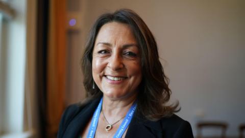 A woman is smiling in a suit. She has brown hair. 