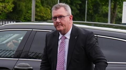 A man with mostly grey hair exiting a car. He is wearing a dark-coloured suit jacket, white shirt and pink tie. He is also wearing glasses. In the background, trees, lampposts and signs are visible