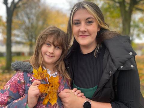 Lisa Bancroft, in a black puffa coat, hugs her daughter Emma, in a grey and pink floral parka, as they hold golden autumn leaves in a park. Trees in autumnal colours are behind them