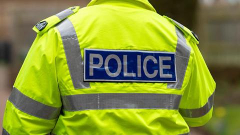 The back of a policeman who is wearing a green jacket with the words "POLICE" written in white font inside a blue rectangle.