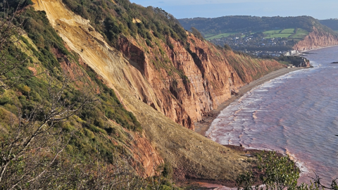 A view of a cliff with a landslide down the middle of it. A village can be seen in the distance behind, with the sea in front to the right and clear blue skies behind.