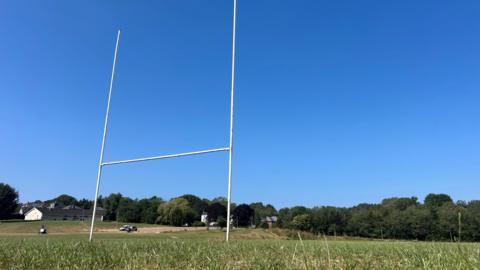 A low down image of rugby posts looking up at them on a clear blue day. The pitch is visible as well as trees in the background.