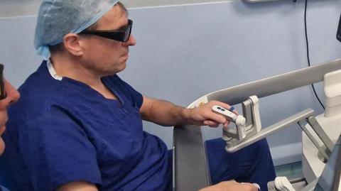 A medic wearing blue surgical scrubs and a head covering is sitting at a screen which shows an operation. He is holding two controls to direct the operation.