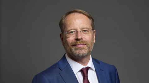 Andy MacNae, wearing glasses and a dark blue shirt and red tie, smiles for the camera in an official photo