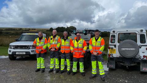 Five men stood shoulder-to-shoulder in front of three 4x4 vehicles. They are all wearing bright yellow and orange, high-viz clothing.