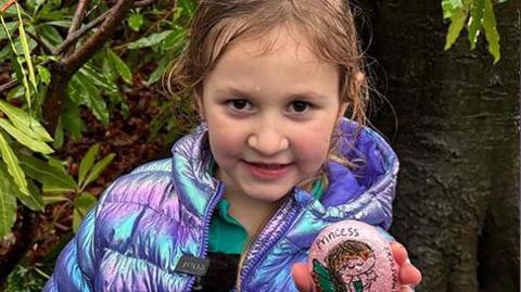 Beatrice, with brown hair and brown eyes, wears a luminous purple and pink coat while holding a rock featuring a painting of a princess. 