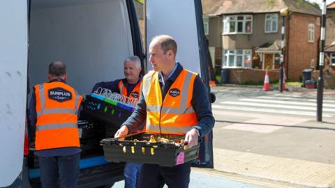 The Prince of Wales carries a crate of food during a public engagement and charity delivery in west London. He wears an orange reflective vest and unloads a white van