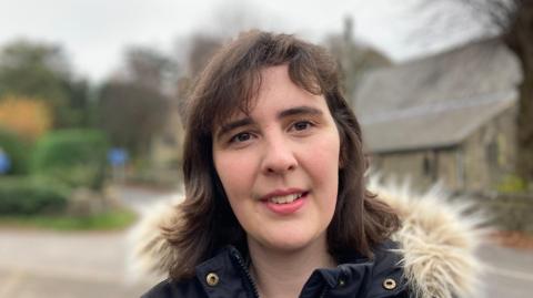 The picture shows Dee Goddard. She is wearing a winter coat and smiling. She is pictured in the Derbyshire Dales village of Curbar