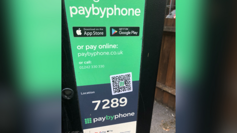 A car parking payment machine with a prominent green paybyphone sign at the centre. A location code is displayed in the middle, but a fake QR code sticker is also placed over part of the sign.