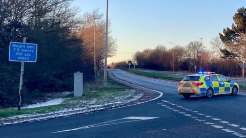 The A120 road which runs through Essex. On the carriageway there is a parked yellow and blue fluorescent marked police car which is blocking the way for any traffic to get down the road. On the right of the picture is a green sign pointing people to Stansted and Braintree.