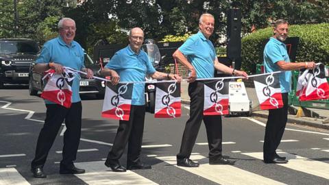 Four members of the choir pose on the famous zebra crossing outside Abbey Road studios. they are all wearing black trousers and light blue polo shirts. They are holding a string of black white and red Black Country flags