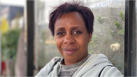 Julie has short dark hair and is half smiling at the camera. She is wearing a grey top and a light grey hoodie over it. In the background is a glass window and a garden.