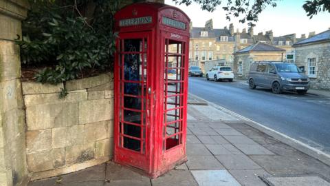A red phonebox on the corner of a road. Cars are parked near by.
